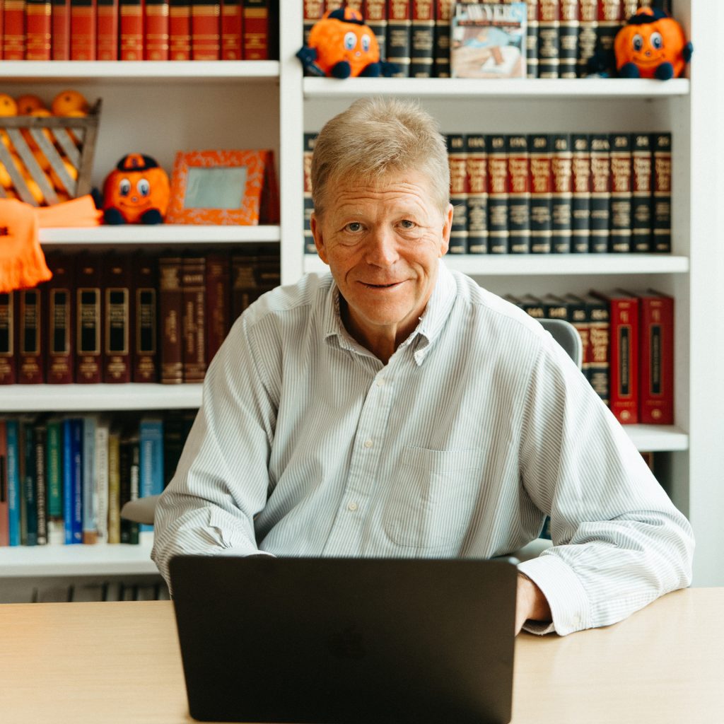 George Boehme looks up from his laptop and smiles. Behind him is a bookshelf.