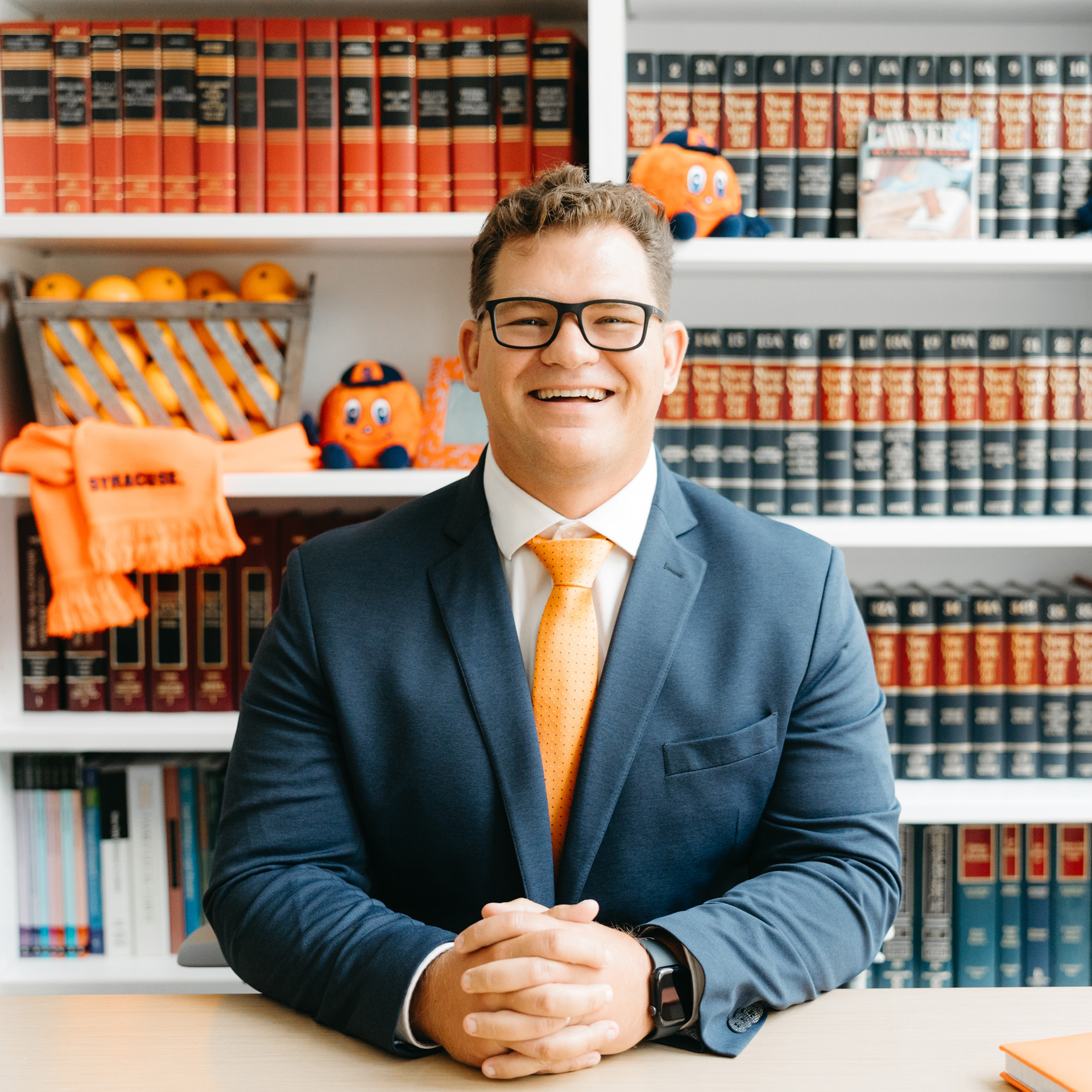 Brette Cox sits in front of book case with orange and blue books with his hands folded at a desk