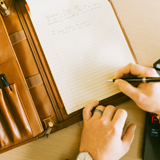 One hand rests on a desk while the other holds a pen over a brown leather padfolio