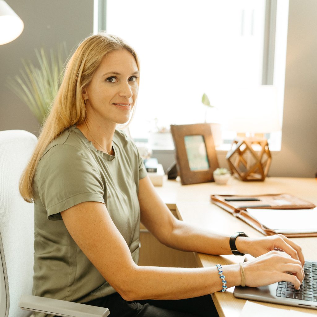 A woman, sitting at a desk in a bright office, looks toward the camera with a soft smile. She is wearing a green blouse and has a laptop in front of her. A Syracuse University pennant hangs on the wall behind her, with plants and office decor completing the background.