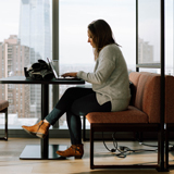 A woman works on a laptop in front of a brightly lit window overlooking a city street